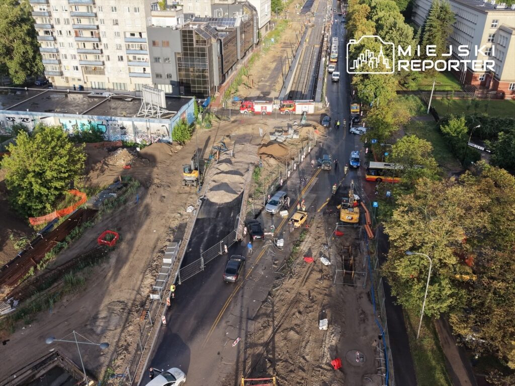 Prace budowlane na ulicy, w tle widoczne budynki mieszkalne i maszyny robocze, a także pojazdy na drodze.