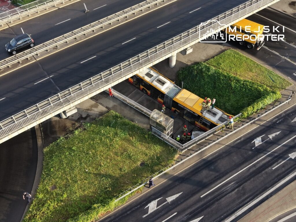 Grupa pracowników w kaskach wykonuje prace konserwacyjne na dachu autobusu pod mostem, w otoczeniu ruchliwej drogi.