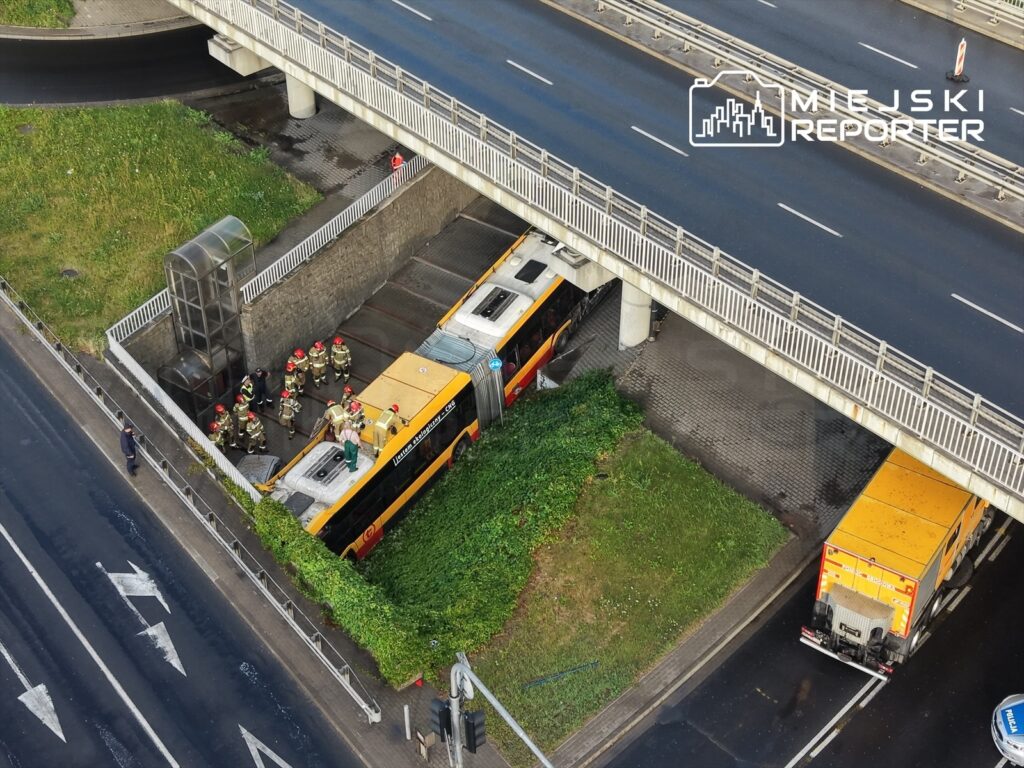 Grupa strażaków w hełmach pracuje przy przewróconym autobusie pod mostem. W tle widoczna droga i pojazd ratunkowy.