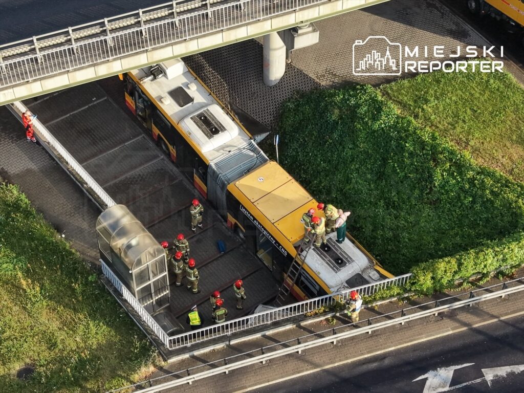 Grupa strażaków w hełmach pracuje przy żółtym tramwaju, który stoi na przystanku pod mostem.