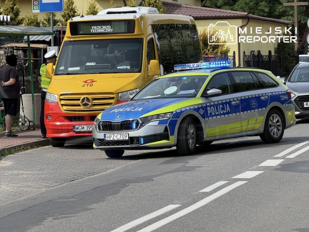 Policjant rozmawia z kierowcą żółtego autobusu, obok stoi radiowóz policyjny z włączonymi sygnałami świetlnymi.