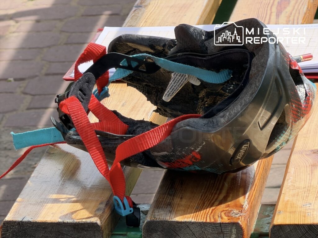 Kask rowerowy z czerwoną i niebieską regulacją leży na drewnianej ławce w parku.