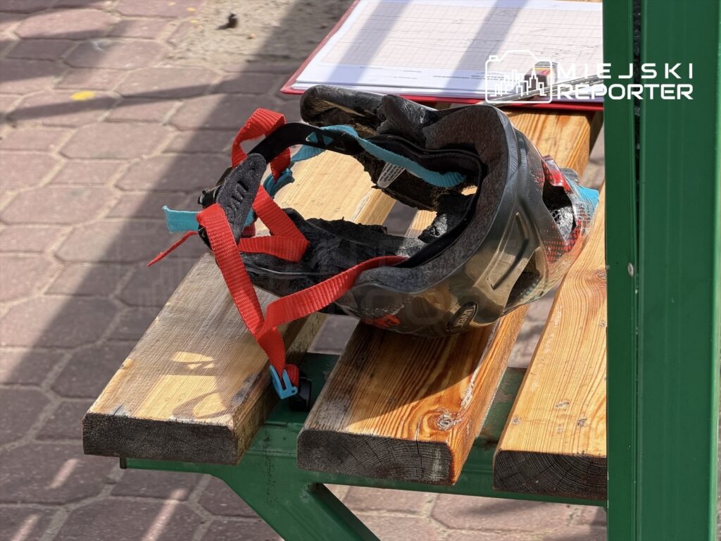 Kask rowerowy z czerwoną i niebieską taśmą leży na drewnianej ławce w słonecznym otoczeniu.