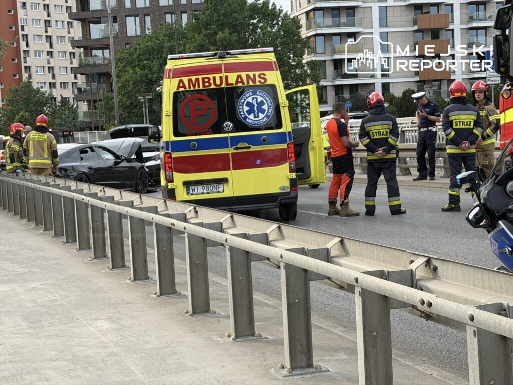 Grupa strażaków i ratowników medycznych z ambulansu ocenia sytuację po zderzeniu dwóch aut na drodze.