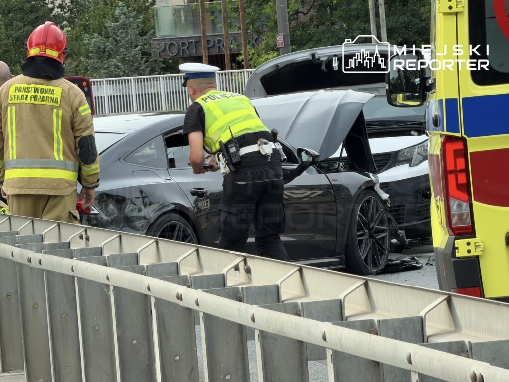 Strażak w mundurze z odblaskami oraz policjant w kamizelce przesłuchują kierowcę przy zderzonych samochodach na drodze.