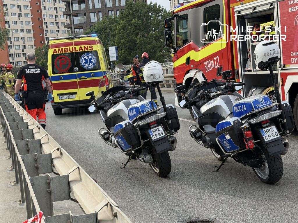 Ratownicy medyczni oraz policja pracują na miejscu wypadku, obok karetki i wozu strażackiego na zablokowanej drodze.