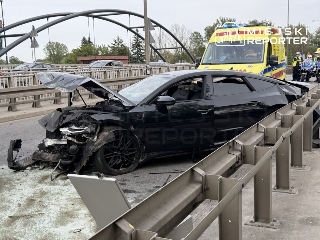 Czarny samochód osobowy z uszkodzoną przednią częścią stoi na jezdni, obok widać ambulans oraz grupę ratowników.