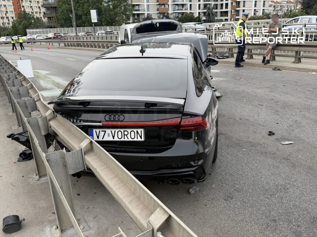 Czarny samochód marki Audi z uszkodzeniami stoi przy barierkach na drodze, obok pracują policjanci.