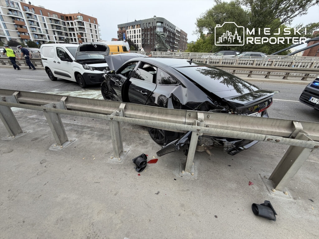 Czarne auto osobowe z uszkodzoną tylną częścią stoi obok białego furgonetki na ruchliwej ulicy.