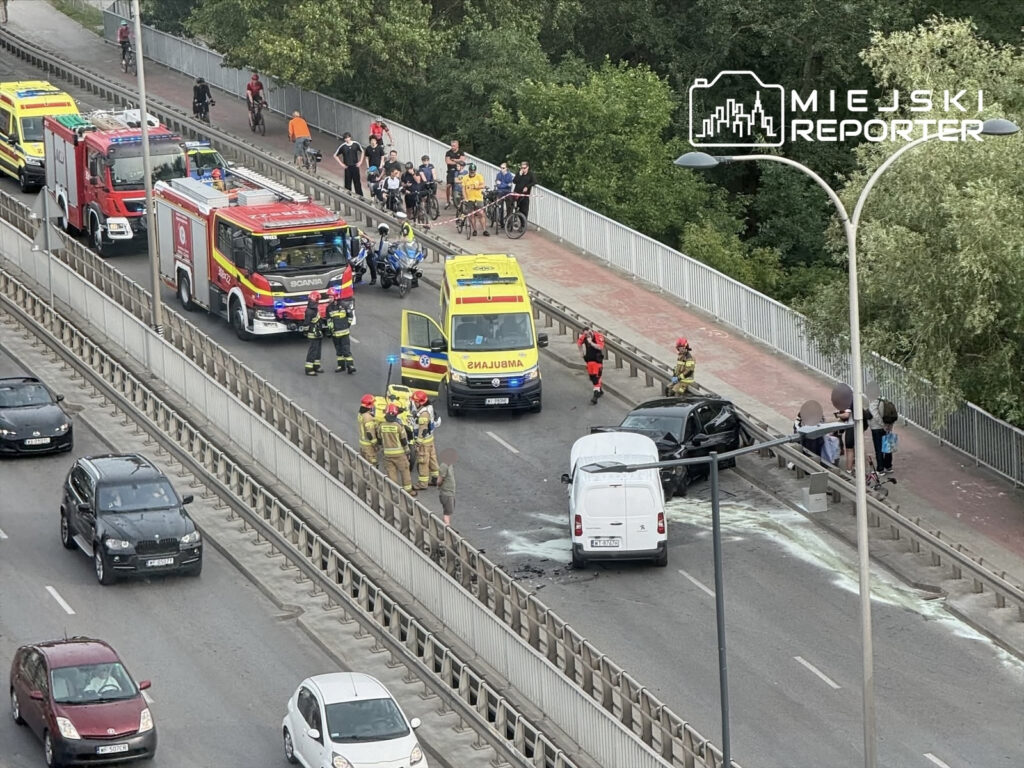 Na drodze widać zderzenie dwóch samochodów, obok pracują strażacy i ratownicy medyczni. W tle grupa osób obserwuje sytuację.