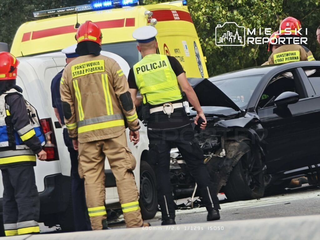 Grupa ratowników i policjantów ocenia uszkodzenia dwóch zderzonych pojazdów na drodze.