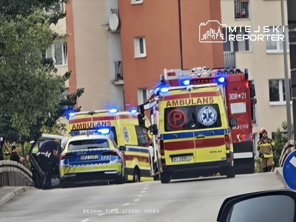 Na ulicy zablokowanej po wypadku stoją karetka pogotowia i radiowóz, obok pracują strażacy w mundurach.