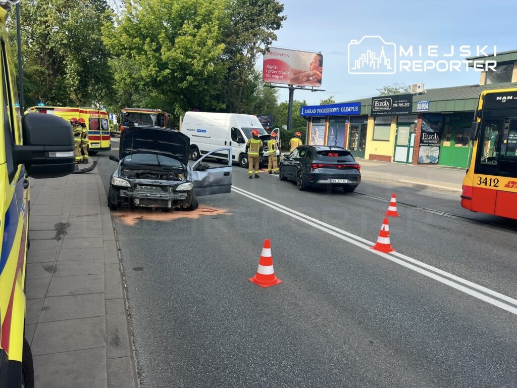 Uszkodzony samochód z otwartą maską na ulicy, wokół niego strażacy i policjanci, w tle cmentarz.