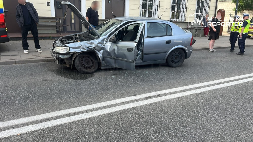 Rozbity samochód osobowy z otwartą maską stoi na ulicy, obok grupka osób oraz radiowóz policji. W tle widoczny cmentarz.