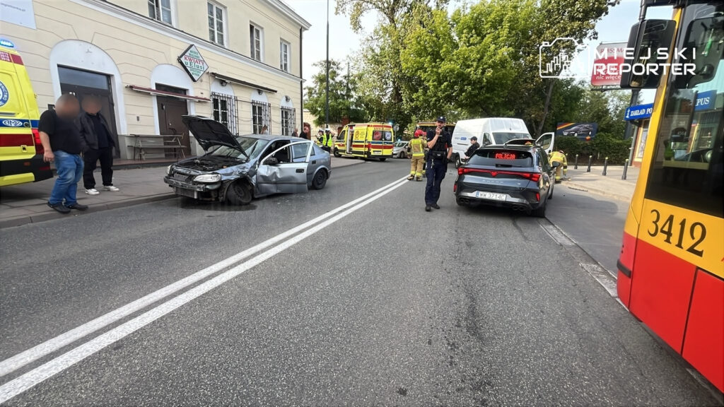 Dwa uszkodzone samochody na ulicy, obok stoi policjant i ratownicy medyczni, w tle widać cmentarz.