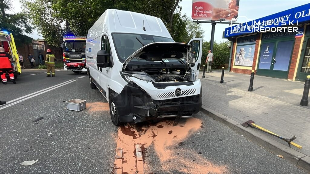 Uszkodzony biały furgon z otwartą maską stoi na ulicy, obok interweniują strażacy i zespół medyczny, a wokół leżą fragmenty szkła.