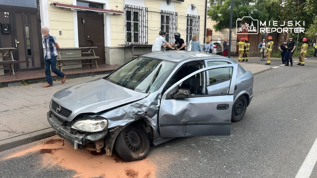 Uszkodzony samochód osobowy stoi na ulicy, obok grupy osób oraz strażaków w mundurach. W tle widoczny cmentarz.