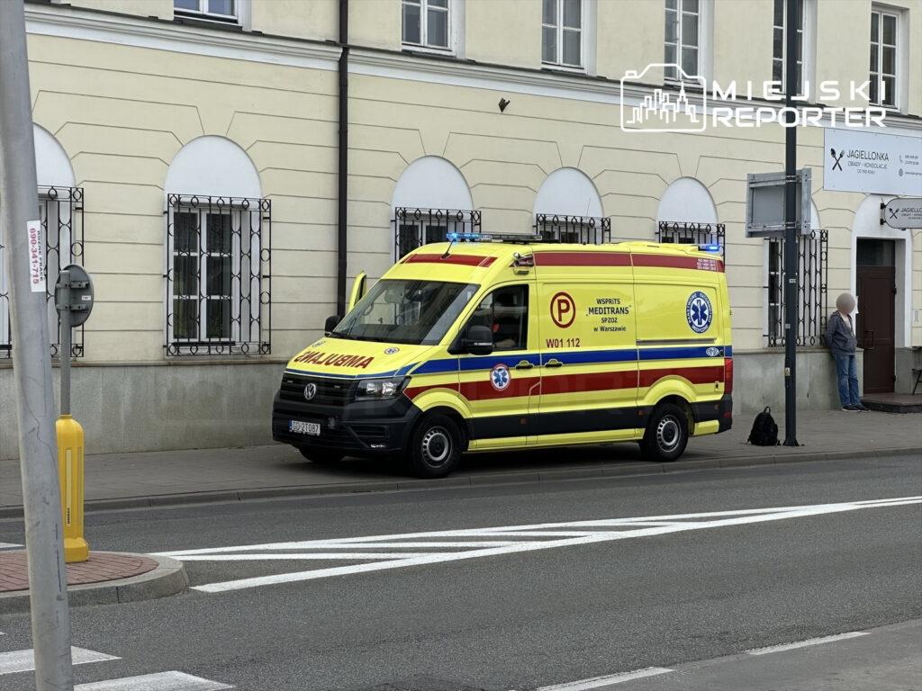 Karetka pogotowia zaparkowana przy chodniku obok budynku, z włączonymi sygnałami świetlnymi.