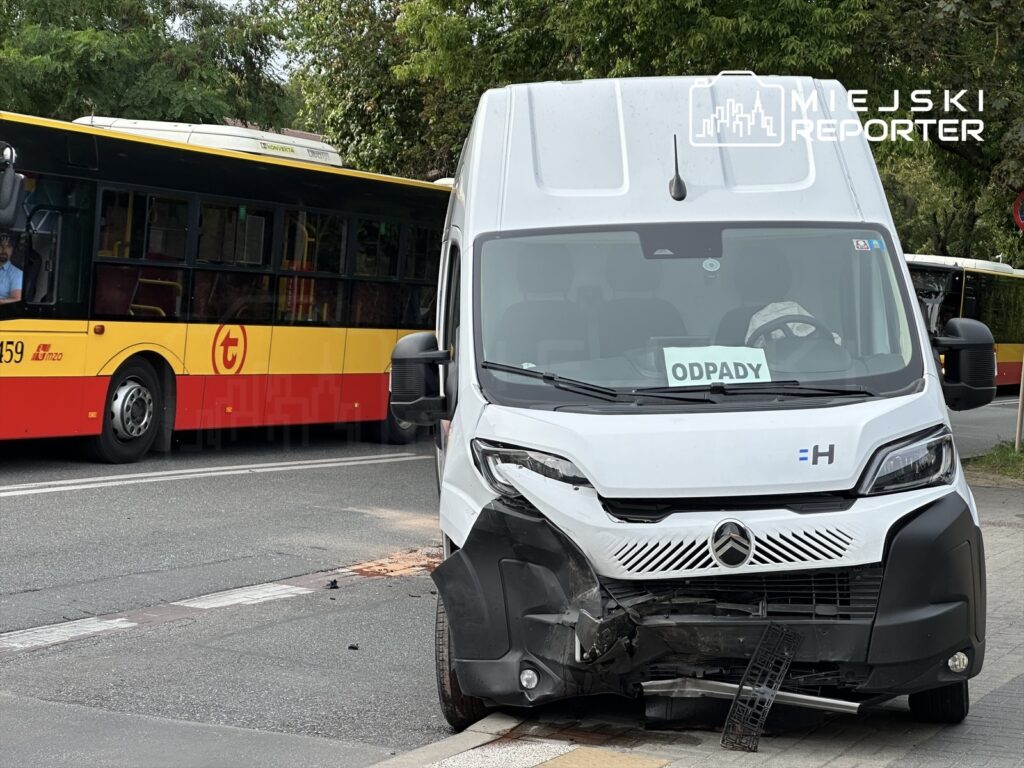 Biały pojazd dostawczy z uszkodzoną przednią częścią stoi na chodniku, obok żółty autobus komunikacji miejskiej. Na szybie pojazdu widoczny jest napis "ODPADY". W tle widać zieleń drzew.