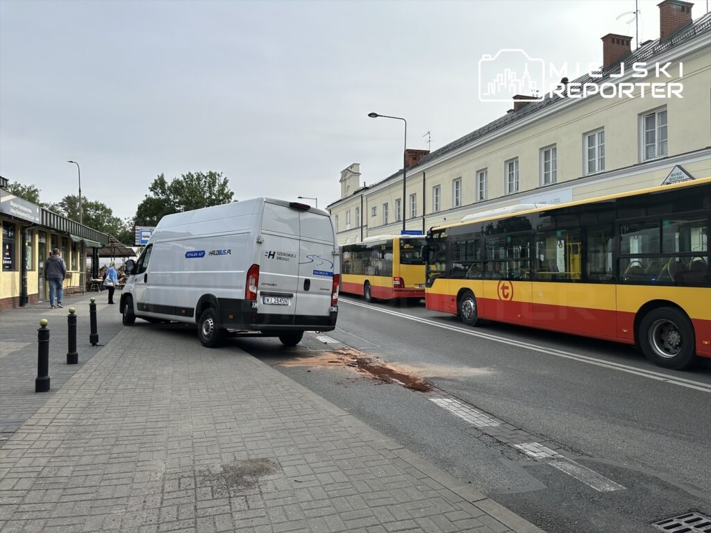 Biały furgon stoi na chodniku obok przystanku, a obok niego przejeżdżają autobusy, w tle widać sklep.