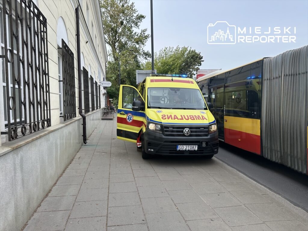 Ambulans z włączonymi sygnałami świetlnymi zatrzymany przy chodniku obok budynku z metalowymi kratami.