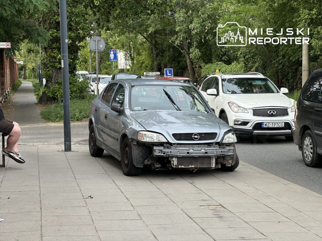 Zniszczony samochód osobowy zaparkowany na chodniku obok ruchliwej ulicy, w tle widoczna zieleń drzew.
