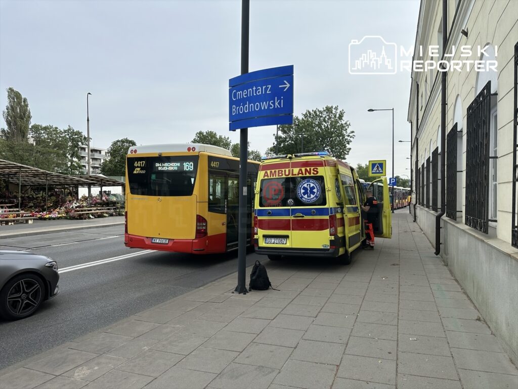 Karetka z włączonymi sygnałami stoi obok przystanku autobusowego, a w tle widać czerwony autobus linii 169. W pobliżu znajduje się znak kierunkowy do cmentarza Bródnowskiego.