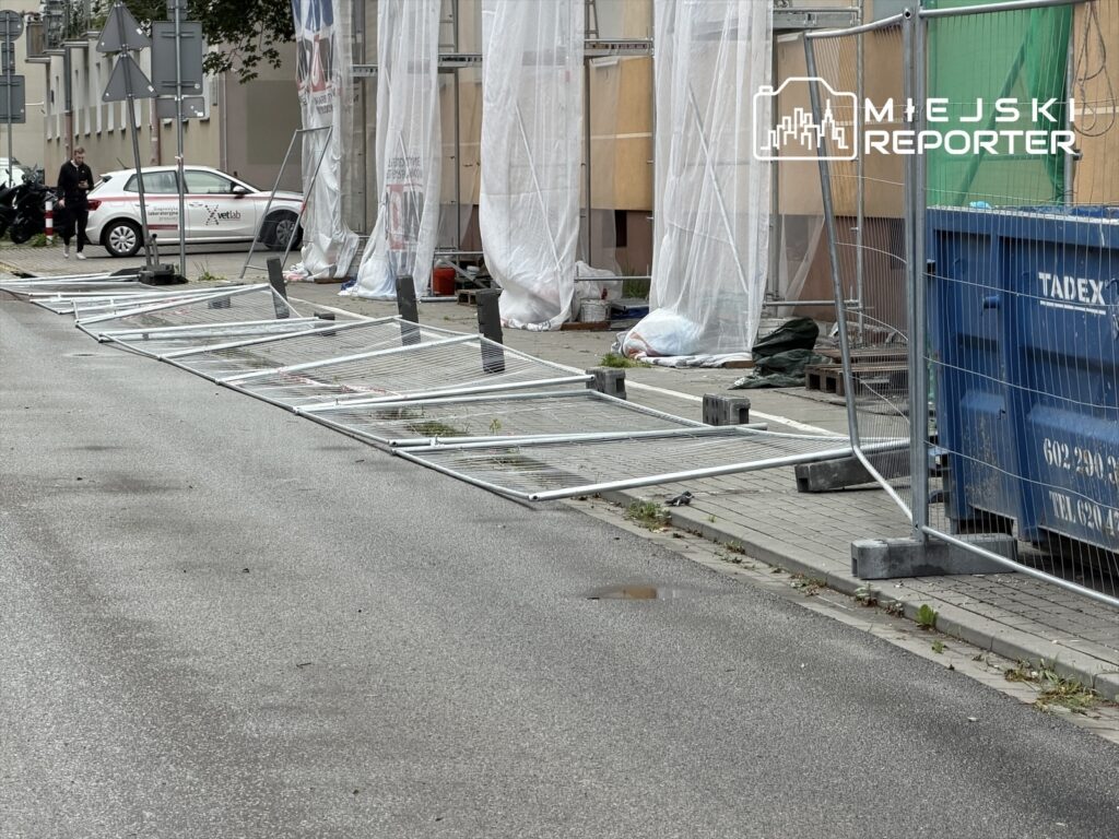 Złamane ogrodzenia leżą na chodniku obok budynku, a w tle widoczny samochód i prace budowlane.