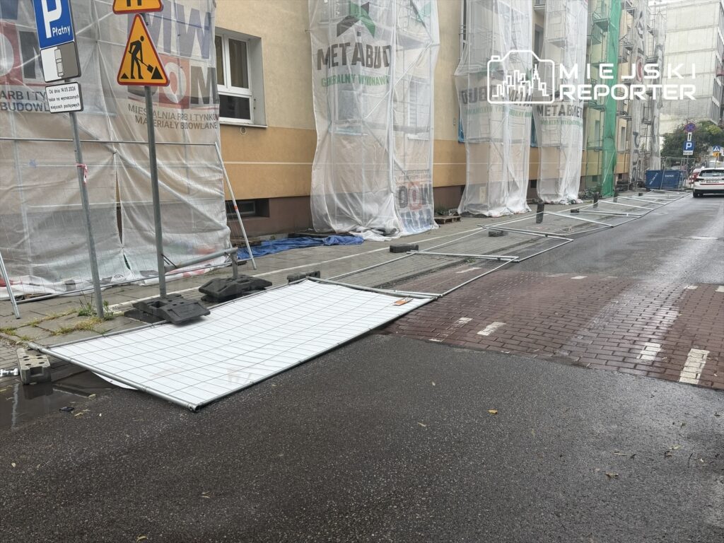 Przewrócone panele ogrodzeniowe leżą na chodniku obok budowy, w otoczeniu plandek i znaków drogowych.