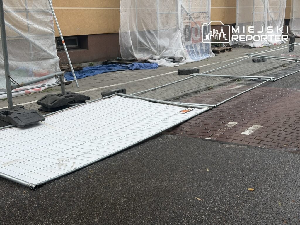 Złamane ogrodzenie budowlane leży na chodniku, a w tle widoczne są plandeki ochronne i puste palety.
