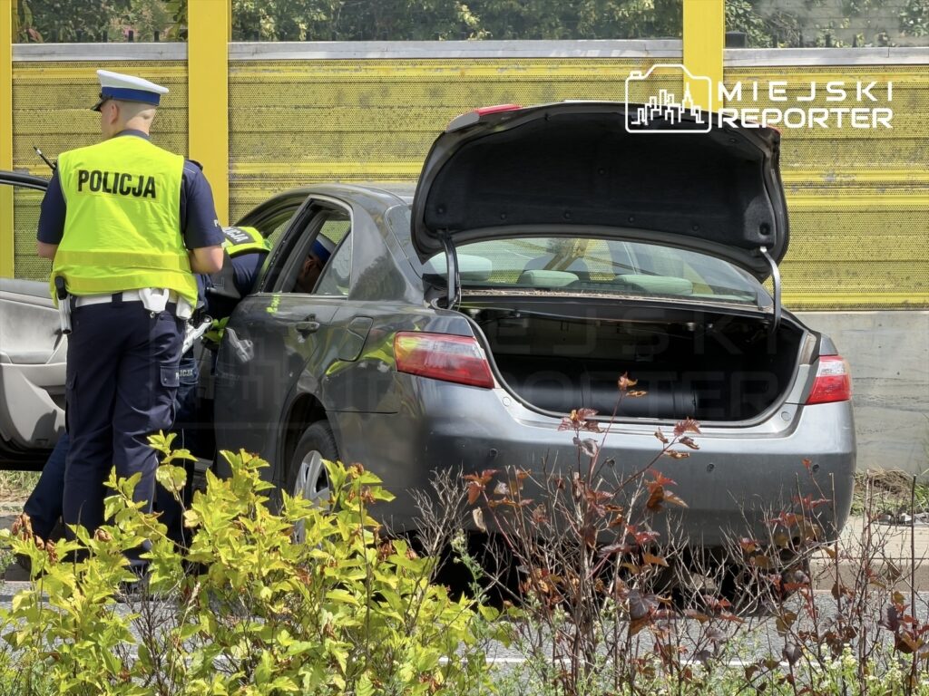 Policjant w kamizelce odblaskowej przeszukuje wnętrze samochodu osobowego na parkingu.