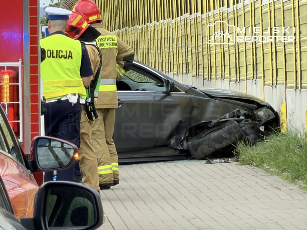 Na miejscu zdarzenia drogowego, ratownicy medyczni i policjanci oceniają uszkodzone auto zatrzymane przy ogrodzeniu.
