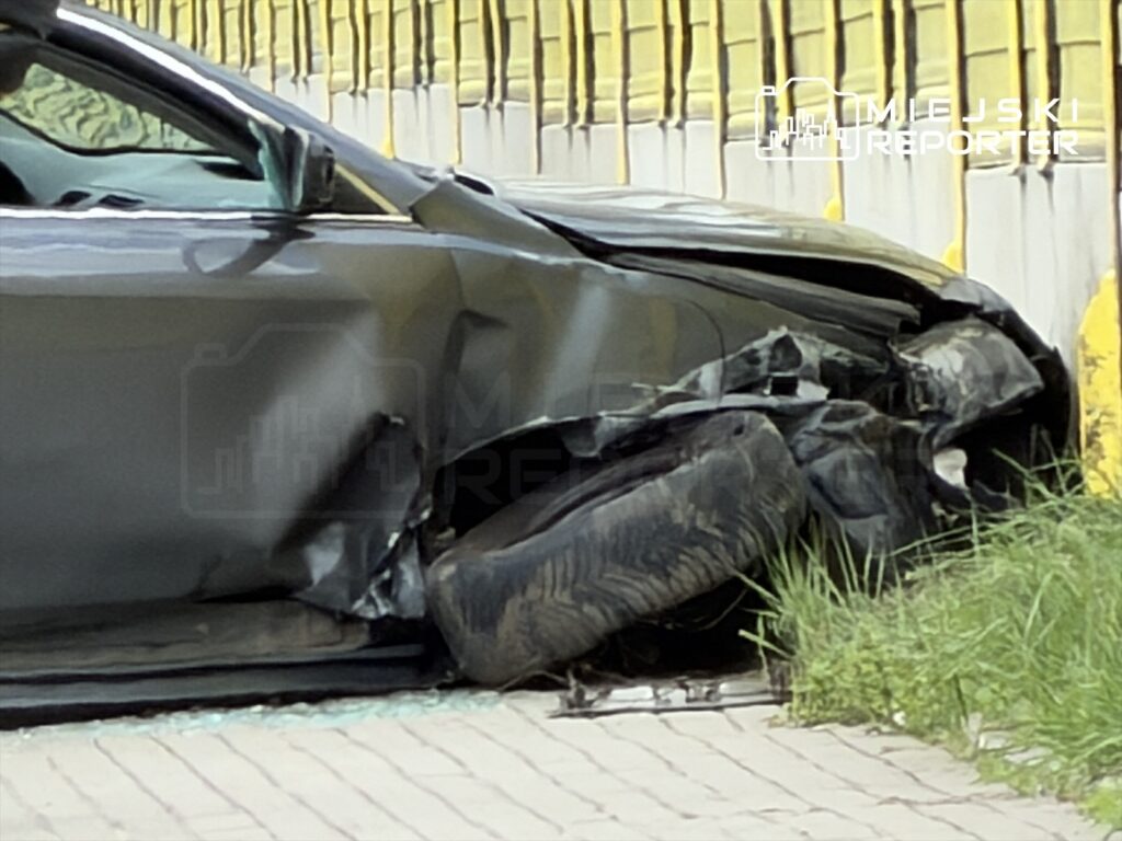 Zniszczony samochód osobowy z uszkodzoną przednią częścią stoi przy ogrodzeniu w zielonym otoczeniu.