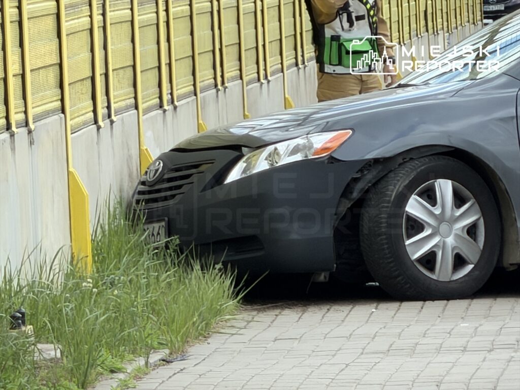 Czarny samochód osobowy zaparkowany blisko ogrodzenia, obok stoi osoba w kamizelce odblaskowej, trzymająca telefon.