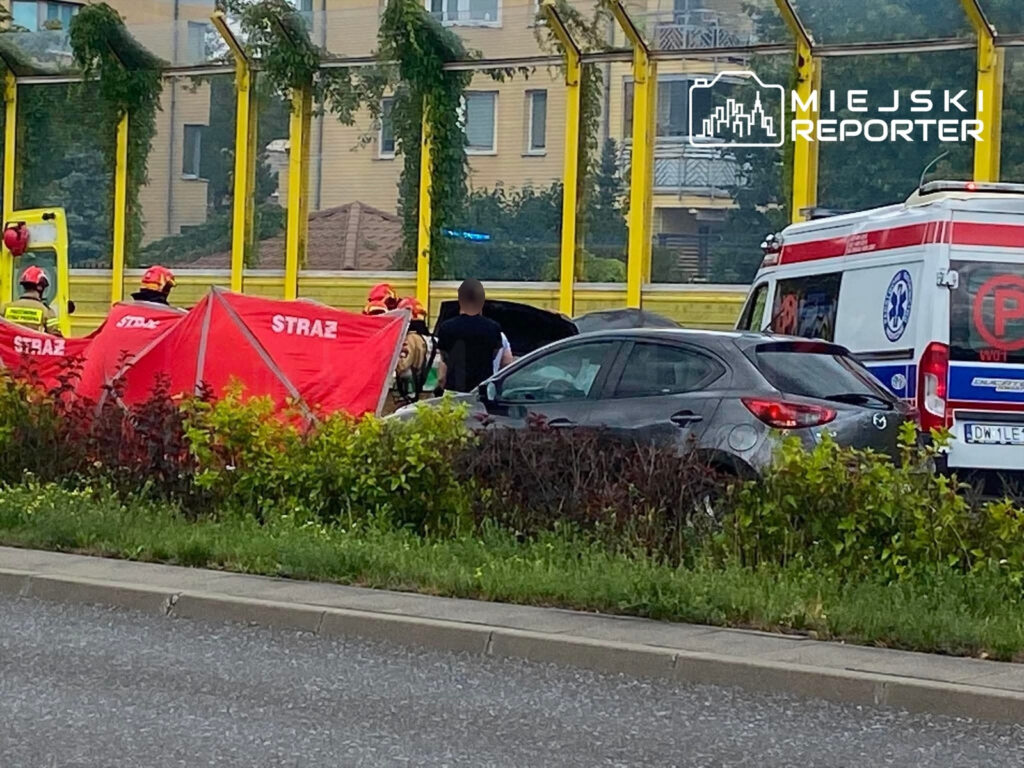 Ratownicy w kaskach pomagają poszkodowanemu przy samochodzie, obok rozstawiony namiot medyczny i karetka.