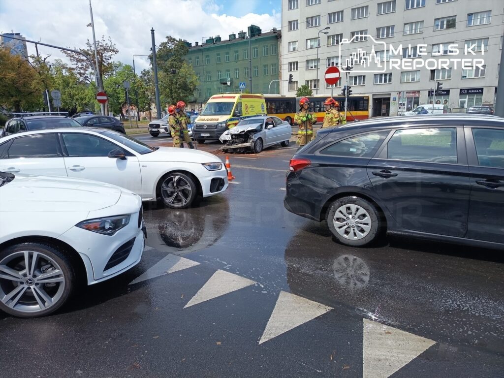 Na skrzyżowaniu kilka samochodów stoi obok siebie, a strażacy w kaskach oceniają zderzenie dźwigu tramwajowego z osobowym BMW.