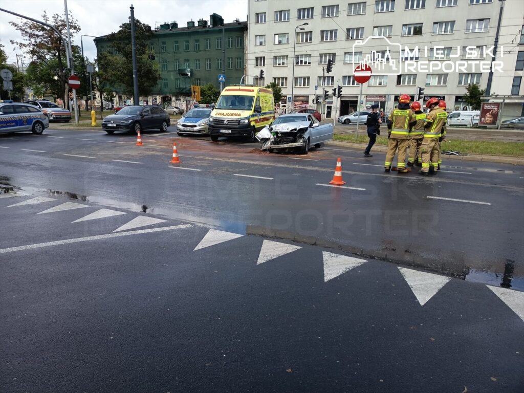 Na skrzyżowaniu stoją strażacy w uniformach, obok rozbitego samochodu osobowego oraz karetki pogotowia.
