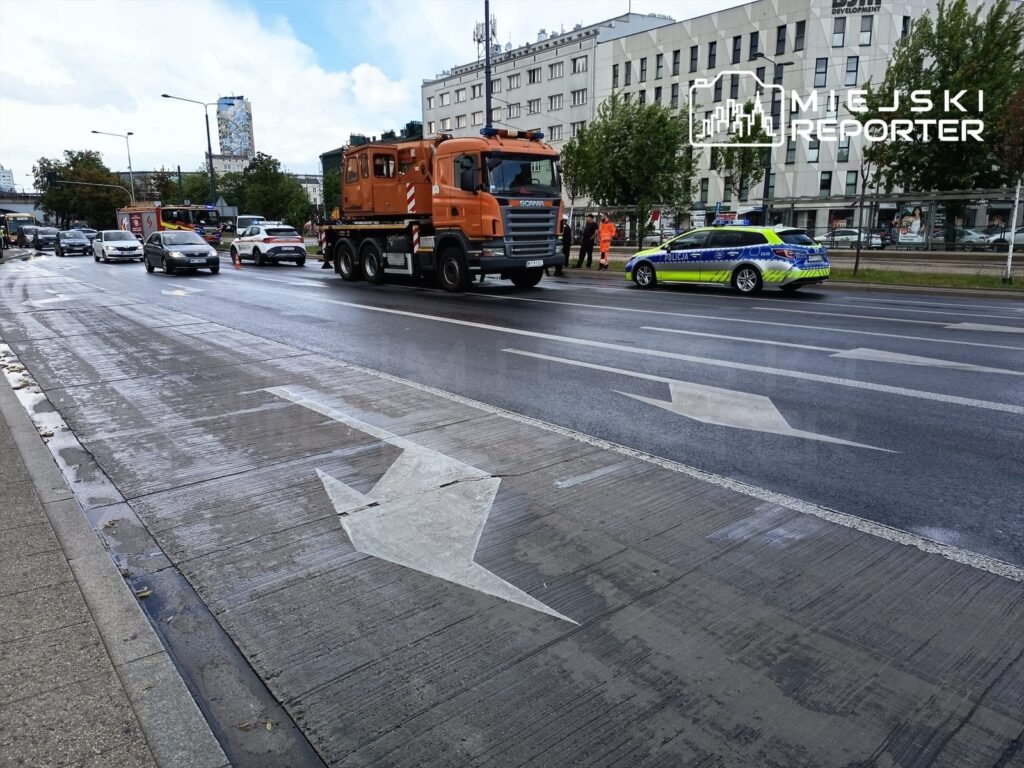 Na jezdni widoczny jest policyjny radiowóz oraz ciężarówka z pomarańczową kabiną, obok stoją samochody osobowe.