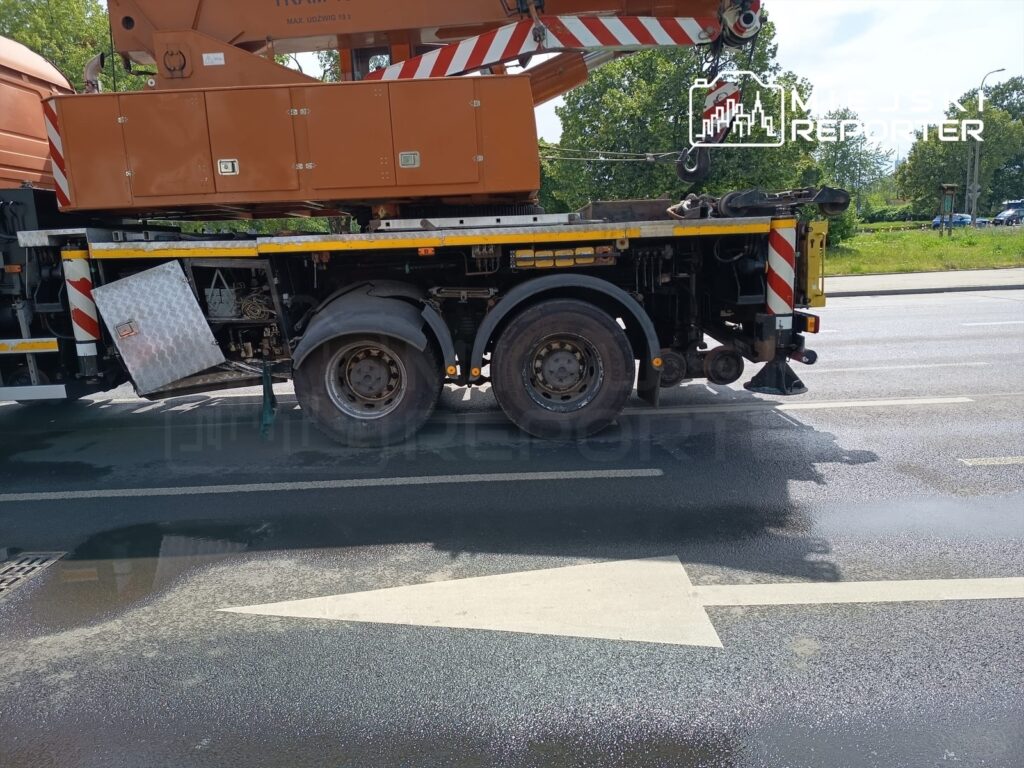 Ciężki dźwig z uniesioną platformą stoi na jezdni, obok widać mokrą nawierzchnię.