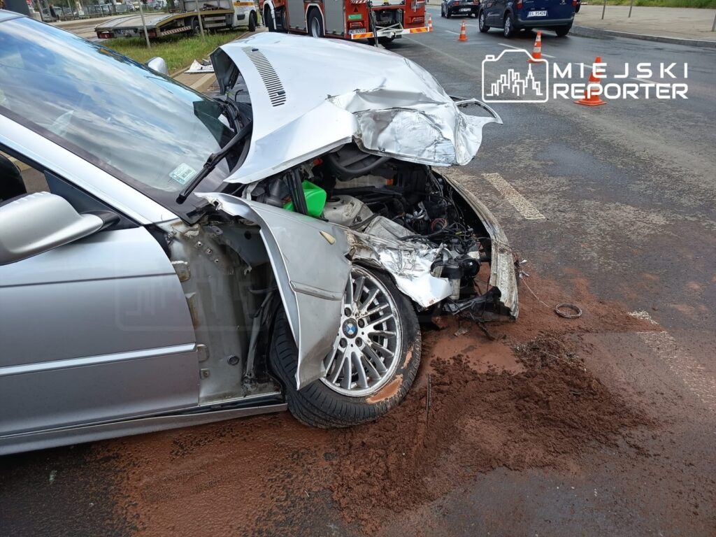 Zniszczony samochód osobowy z rozbitą przednią częścią leży na drodze, obok widać ślady ziemi i pojazdy służb ratunkowych.