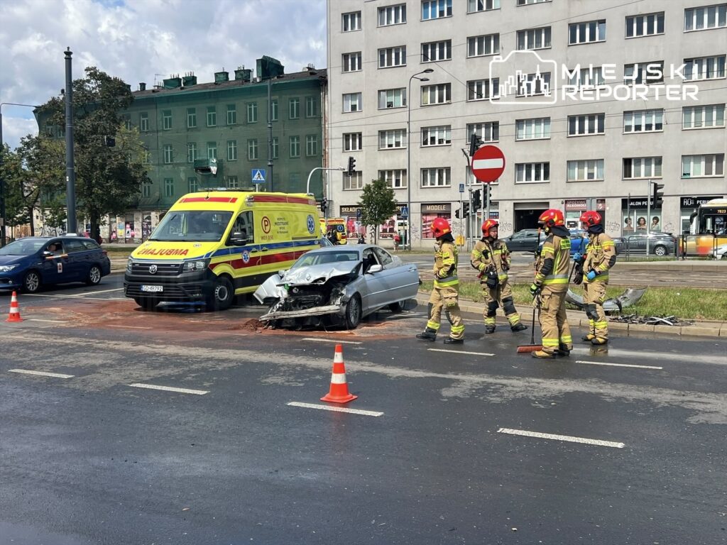 Ratownicy w mundurach omawiają sytuację obok uszkodzonego samochodu na ruchliwej ulicy, gdzie stoi karetka.