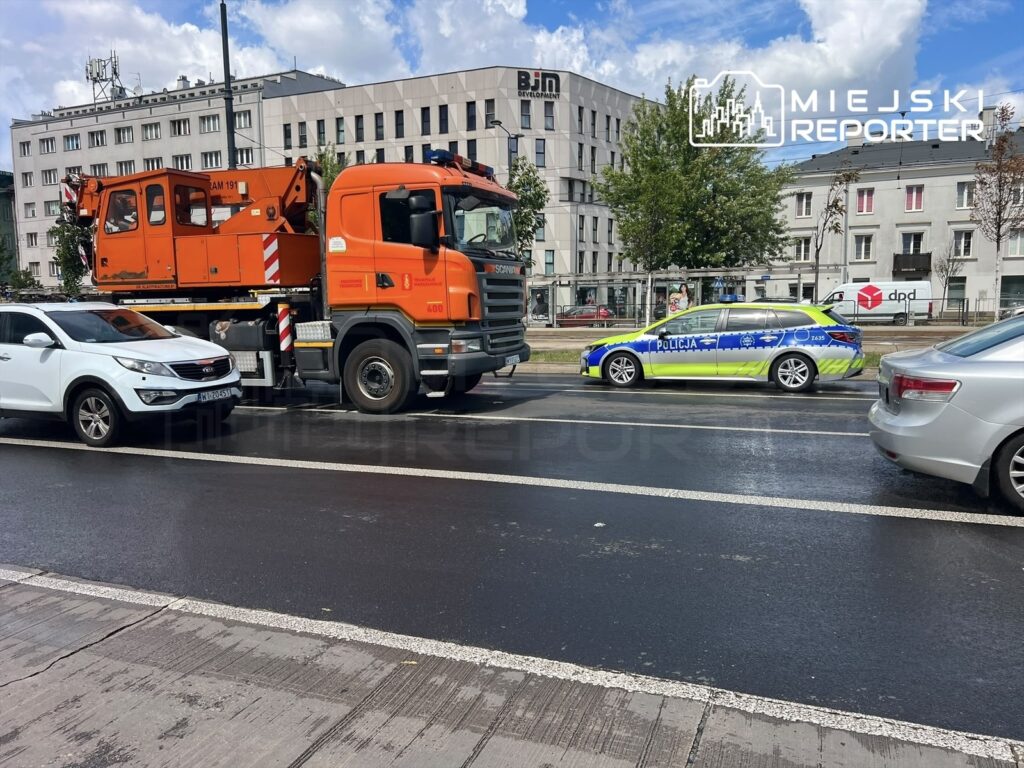 Pomarańczowy dźwig tramwajowy stoi obok białego samochodu osobowego, a obok niego zaparkowany radiowóz policji na ulicy.