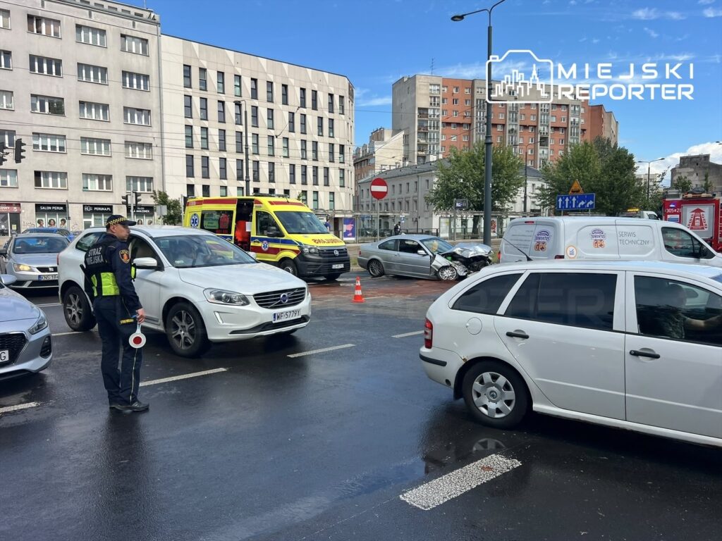 Policjant kierujący ruchem stoi obok uszkodzonego samochodu na skrzyżowaniu, obok widać karetkę pogotowia.