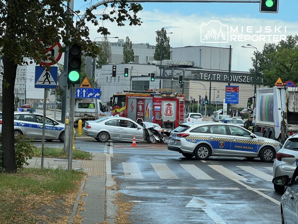 W miejscu wypadku na skrzyżowaniu, uszkodzony samochód osobowy stoi obok dźwigu tramwajowego, a w tle widoczne są pojazdy służb ratunkowych.