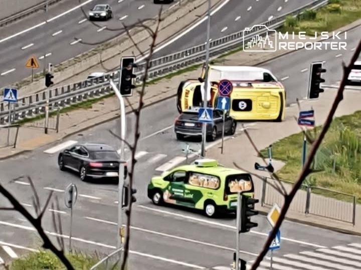 Czarny samochód osobowy zatrzymany na skrzyżowaniu, obok przewrócony żółty pojazd dostawczy oraz zielony samochód z reklamą.