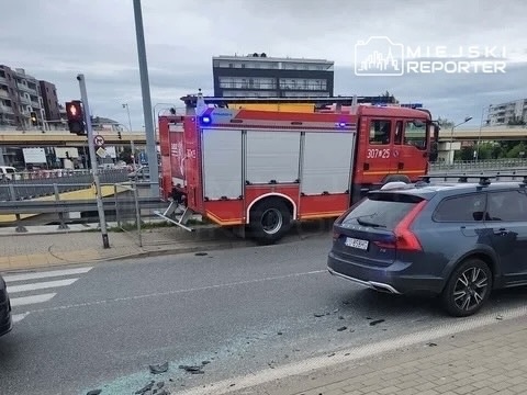 Czerwony wóz strażacki z włączonymi sygnalizatorami zatrzymany na ulicy obok uszkodzonego samochodu.