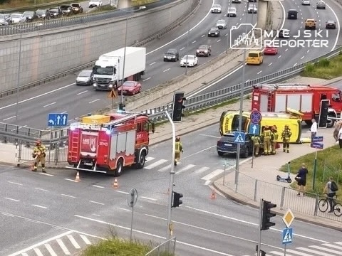 Na skrzyżowaniu dróg stoją dwa wozy strażackie, a obok nich pracują strażacy przy przewróconym pojeździe.