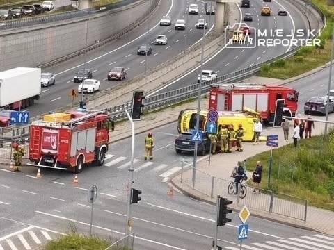 Strażacy interweniują przy przewróconym samochodzie na skrzyżowaniu, obok stojące pojazdy ratunkowe.