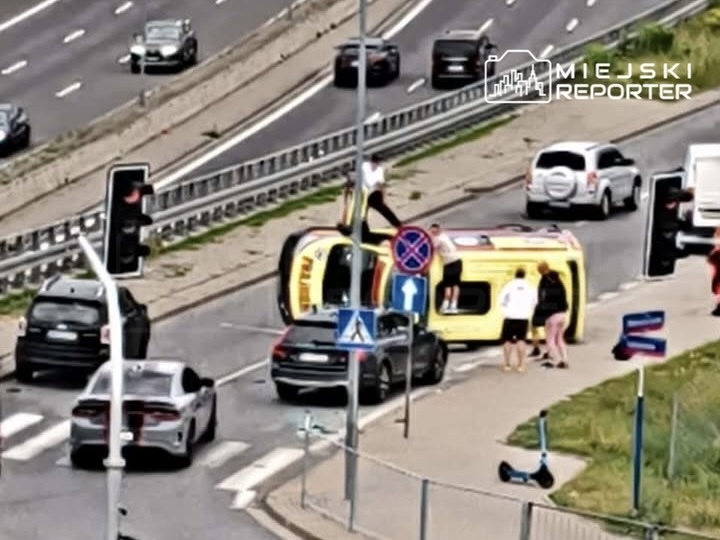 Na drodze leży przewrócony żółty autobus, a obok niego grupka osób interweniuje, podczas gdy inne pojazdy czekają w korku.