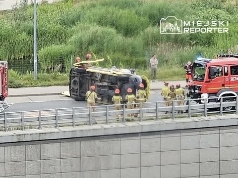 Grupa strażaków w hełmach otacza przewrócony samochód na drodze, obok stoi wóz strażacki.